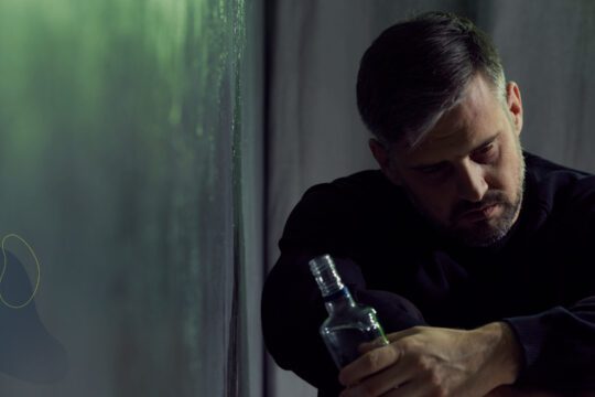 Mężczyzna siedzący w półcieniu trzyma butelkę alkoholu, symbol wewnętrznego napięcia i poszukiwania ulgi